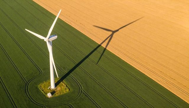 Aerial wind turbine casting long shadow across green crop field and golden harvest