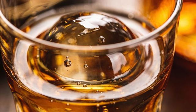 Macro bourbon in crystal tumbler with large ice sphere and amber backlit luxury bokeh