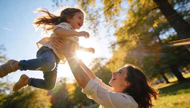Mother lifting laughing daughter during golden hour park play with sun flare and joyful family bond