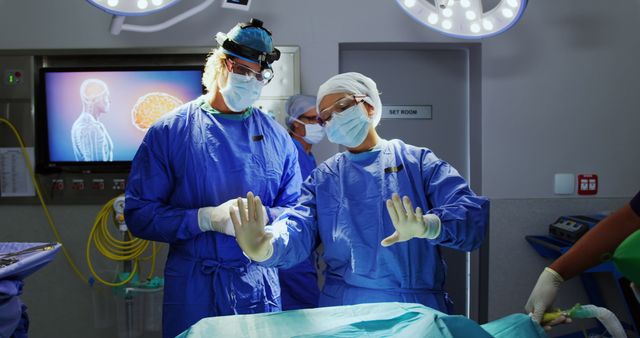 Two surgeons wearing blue surgical attire and masks preparing for brain surgery. They are adjusting sterile gloves in a brightly lit operating room with specialized surgical equipment and displays visible. This image can be used for promoting healthcare services, medical education materials, articles on surgical procedures, or depicting teamwork and professionalism in the medical field.
