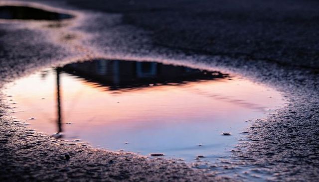 Pastel sunset reflecting in puddle on wet asphalt with roofline silhouette