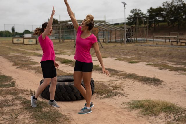 Two Caucasian women wearing pink t shirts at a boot camp training session, exercising, celebrating and high fiving with tyre next to them. Outdoor exercise, fun healthy challenge.