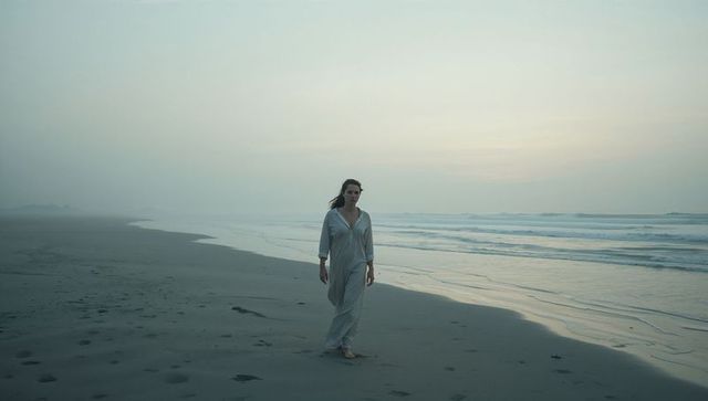 Solitary barefoot woman walking along misty low-tide beach leaving footprints in wet sand
