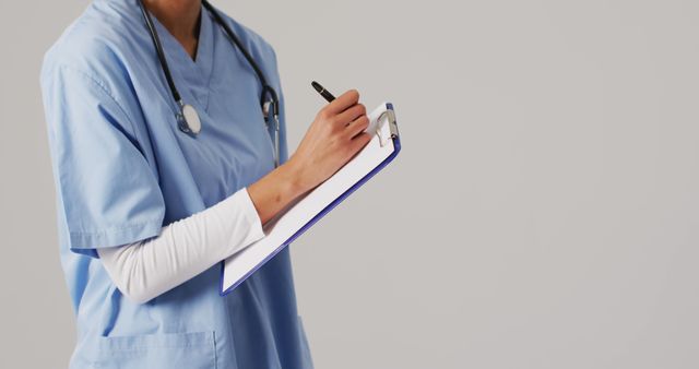 Healthcare professional in blue uniform writing on clipboard with stethoscope around neck. Useful for medical, healthcare, hospital, nursing, and doctor-related topics.