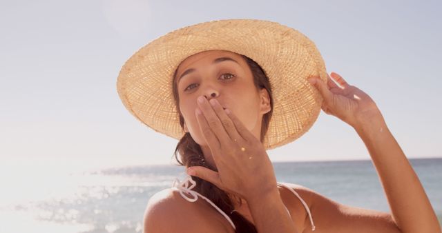 A young woman enjoys a sunny beach day, wearing a straw hat and sending a kiss, with copy space. Her relaxed demeanor and seaside attire suggest a leisurely vacation vibe.