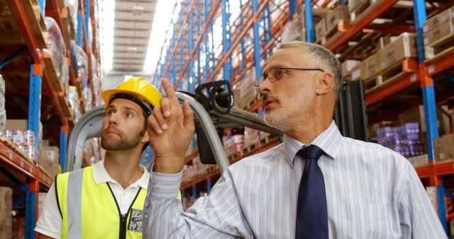 Warehouse supervisor giving instructions to a worker in a safety vest and hard hat amidst rows of inventory. Ideal for use in topics related to logistic operations, industrial management, workplace safety, and inventory control.