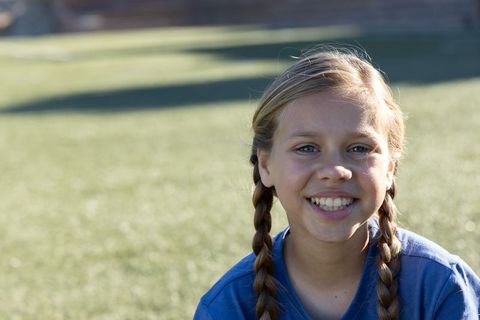 Smiling girl with braids on sunlit grassy field, casual blue tee, outdoor youth portrait