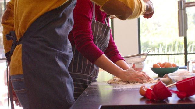 Diverse couple enjoying time together making dough in brightly lit kitchen. Ideal for concepts of togetherness, culinary skills, happiness in domestic life, and baking tutorials. Useful in home cooking blogs, lifestyle articles, and cooking product advertisements.