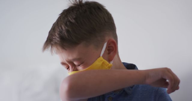 A young boy wearing a yellow face mask coughs into his elbow, demonstrating proper hygiene and illness prevention. This image may be used in health education materials, public health campaigns, or articles about virus prevention and hygiene practices.