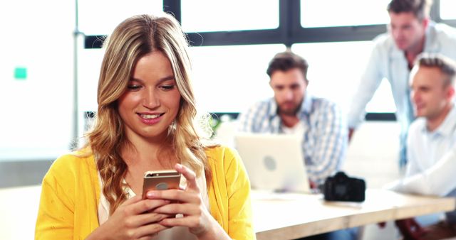 This image captures a young woman engaging with her smartphone in a contemporary office environment while her colleagues work together in the background. The scene exudes a sense of casual yet professional interaction, making it ideal for use in business marketing, team-building promotions, or content related to modern work practices and technology integration.