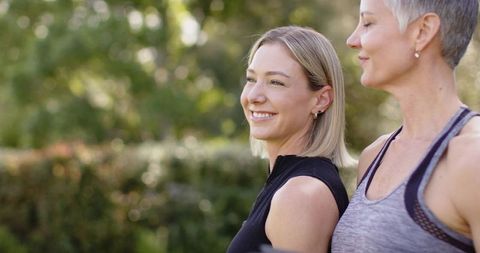 Two women smiling on sunny park walk wearing casual activewear, hoop earrings, soft bokeh