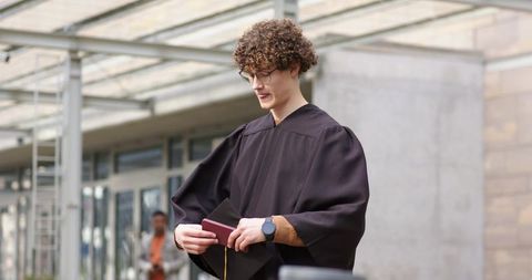 Young graduate in dark gown and glasses holding diploma cover under modern glass canopy