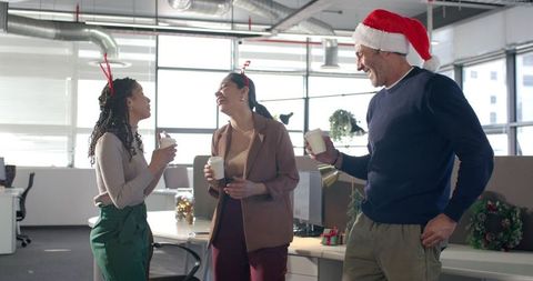 Multicultural coworkers laughing and holding coffee during festive office holiday party