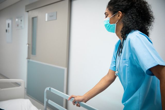 This image shows a female doctor wearing a face mask and pushing a patient's bed in a hospital corridor. It is ideal for use in articles, blogs, and websites related to healthcare, medical services, hospital care, and COVID-19 safety measures. It can also be used in educational materials and promotional content for healthcare facilities.