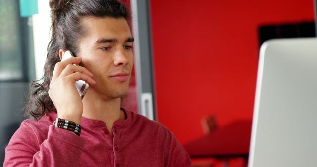 Young man having a phone conversation while seated at a desk with a computer. Ideal for use in business, technology, and communication themes to illustrate remote work, multitasking, or modern office environments.