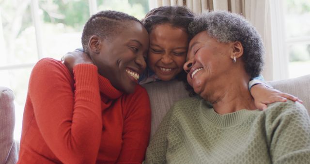 Three women of different generations, including a grandmother, a mother, and a daughter, are bonding at home while sitting on a couch and embracing each other. They are smiling warmly, reflecting a moment of family unity and love. The image is perfect for content related to family values, generational experiences, and home life.