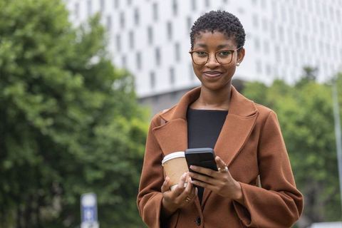 Confident african businesswoman holding coffee and smartphone beside modern office plaza