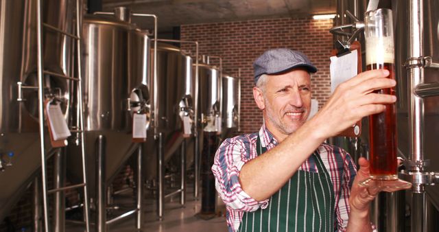 Mature man wearing checkered shirt and striped apron inspecting tall glass of beer in microbrewery. Industrial brewing tanks visible in background, brick walls adding to classic brewery feeling. Useful for illustrating craft beer industry, brewing process, small business, and beer quality control methods.
