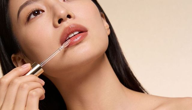 Asian woman applying glossy lip gloss with gold wand, dewy makeup close-up
