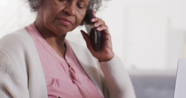 This image depicts a senior woman engaged in conversation on her phone while typing or working on a laptop at home. Useful for illustrating themes related to senior citizens, technology usage among elderly, modern communication, or at-home activities. Ideal for use in blogs, articles, and advertisements focused on elderly care, digital technology integration, or lifestyle of older adults.