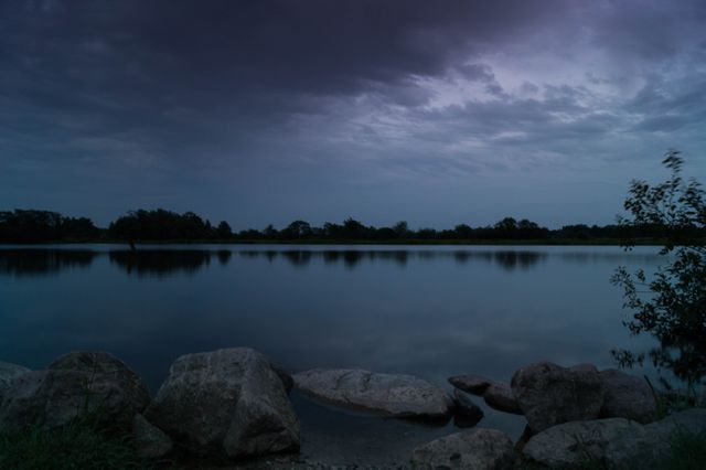 Photo captures a serene lake at dusk with a dramatic, overcast sky reflecting in the calm waters. Features large rocks in the foreground and distant trees. Ideal for use in nature-inspired projects, relaxation content, or atmospheric landscape scenes.