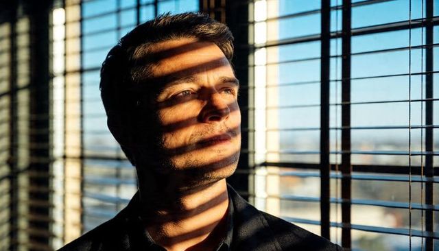 Cinematic contemplative man gazing through blinds with dramatic striped sunlight