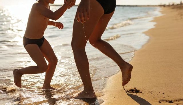 Parent and child running hand-in-hand on sunlit beach at golden hour, splashing surf