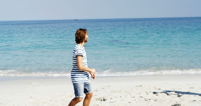 Man dressed in striped shirt and shorts walking along a sandy beach with a clear blue sea in the background at daytime. Perfect for illustrating vacations, leisure activities, relaxation, coastal getaways, and beachside retreats.