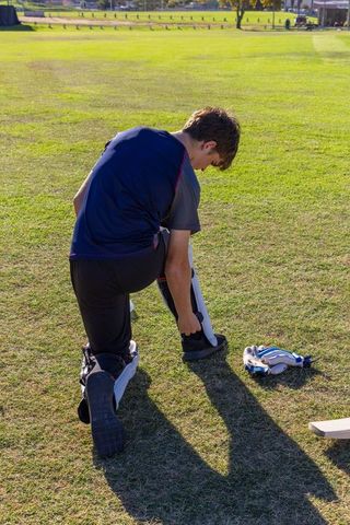 Male cricketer kneeling fastening leg pads on sunlit grassy sports field preparing for match