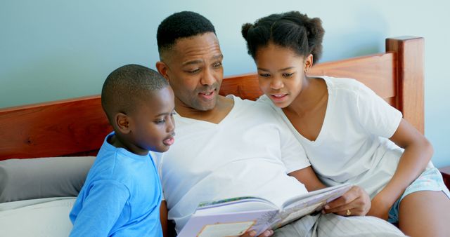Front view of mid adult black father with his children reading story book in bedroom of comfortable home. They are relaxing on bed 4k