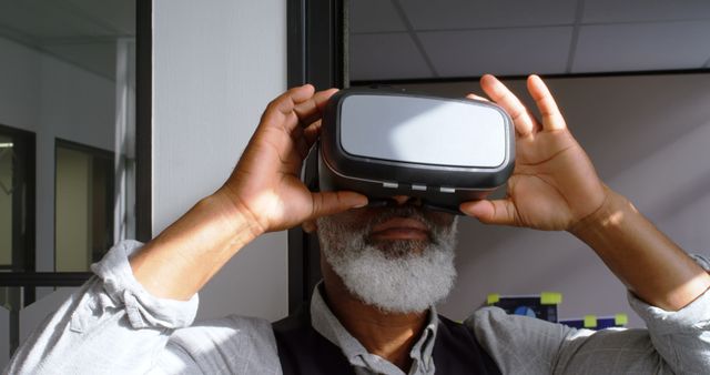 Senior man exploring the digital world wearing a VR headset indoors. Ideal for technology innovation, modern lifestyle, and digital experience concepts. Useful for articles or advertisements related to virtual reality, seniors embracing new technology, and futuristic experiences.