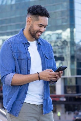 Smiling young man using smartphone outside modern glass office building commuter lifestyle