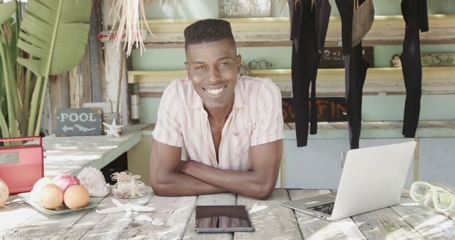 Portrait of happy african american man in surf shop on beach with laptop and tablet. Lifestyle, business, vacation, summer and leisure, unaltered.