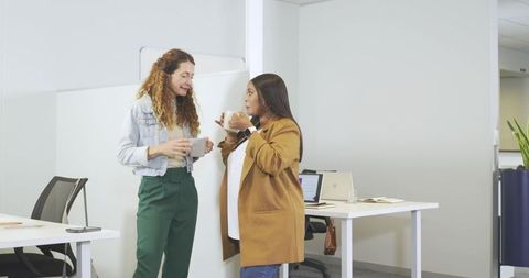 Diverse female coworkers chatting over coffee holding mugs in modern minimalist office