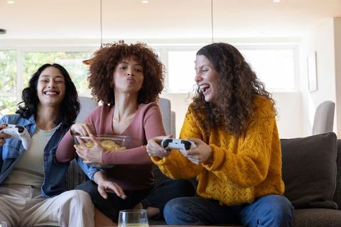 Diverse women playing video games on sofa laughing and sharing snacks at home