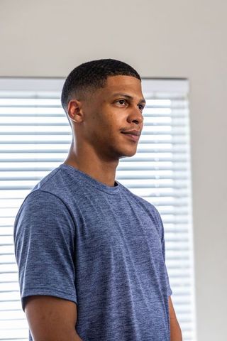 Young man standing by sunlit blinds wearing blue t-shirt looking thoughtful minimalist portrait