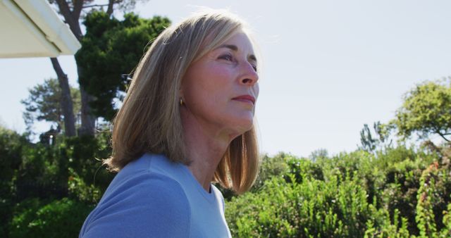 A mature woman with blonde hair, dressed in a light blue top, contemplates while enjoying a sunny day outdoors surrounded by lush greenery. Ideal for use in articles related to aging gracefully, retirement lifestyle, mental wellness for seniors, serene outdoor escapes, or personal reflections.
