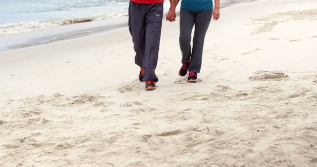 Couple walking on sandy beach holding hands, embodying love and companionship. Might be used for advertisements for travel destinations, romantic getaways, wellness and fitness promotions or inspirational blog posts about health and relationships.