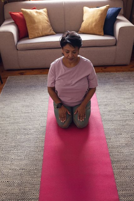 High angle view of biracial mature woman with eyes closed kneeling and meditating on mat at home. Unaltered, exercise, yoga, retirement, zen, healthy and active lifestyle concept.