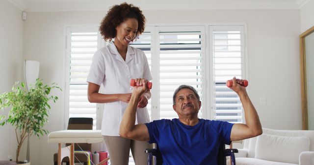 Elderly man using hand weights for exercising while seated in a wheelchair, assisted by a smiling physical therapist. Ideal for content related to senior care, rehabilitation therapy, healthcare, and home exercise programs.