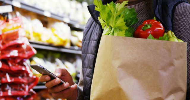 Close-up of person shopping in supermarket, holding a paper bag with fresh vegetables and using a smartphone. This image can be used in articles or advertisements about grocery shopping, healthy eating, supermarket sales, and other lifestyle or technology-related content.