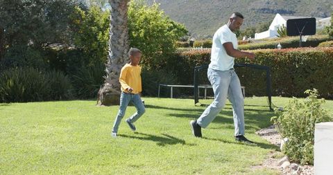 African american father and son walking on sunlit lawn playing basketball in yard