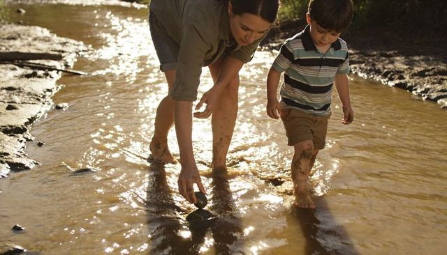 Mother and young son wading muddy creek at golden hour picking stones and exploring