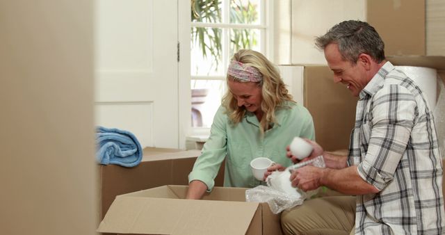 A middle-aged Caucasian man and woman are unpacking items from a box, with copy space. Their smiles suggest a positive new beginning, moving into a new home.