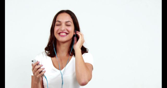 Young woman smiling and enjoying music while using blue earphones and holding a smartphone. Person is wearing a white shirt and standing against a plain white background, portraying joy and relaxation. Ideal for advertisements, social media content, health and wellness websites, or promoting music apps and audio products.