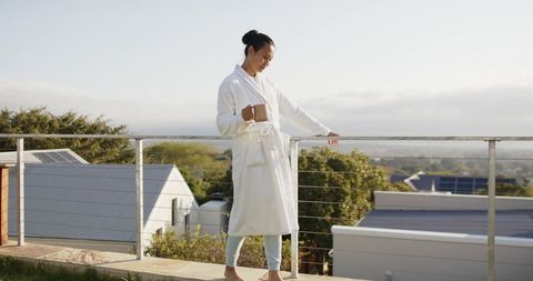 Asian woman enjoying sunrise coffee on balcony in white bathrobe overlooking suburban view