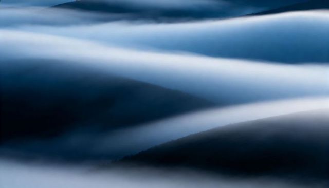 Flowing mist over rolling mountain ridges at dawn creating layered blue abstract landscape