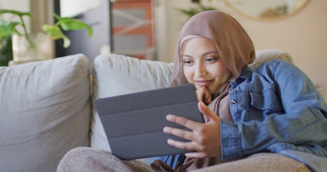 Young Muslim woman relaxing on a couch and using a digital tablet at home. She is wearing a hijab and casual clothes, sitting comfortably and looking at the screen with a smile. Ideal for themes related to technology in daily life, home comfort, digital media consumption, and the lifestyle of Muslim women.