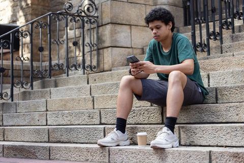 African american man sitting on urban stone steps checking smartphone with coffee cup