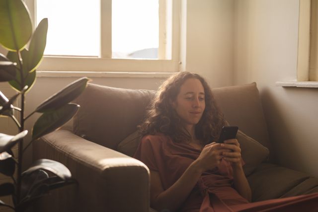 This image depicts a woman sitting comfortably on a sofa, using her smartphone. The natural light from the window creates a warm and cozy atmosphere. This image can be used for articles or advertisements related to home lifestyle, technology use, quarantine activities, or promoting relaxation and comfort at home.